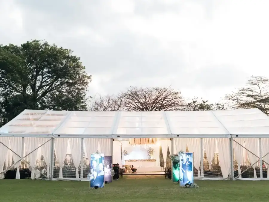 Exterior evening view of a large clear-span marquee tent with white drapes and warm lighting, set