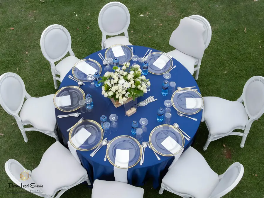 Aerial view of a round table set up with a navy blue tablecloth, gold-rimmed clear glass chargers, white napkins, wine glasses, and a white floral centerpiece.
