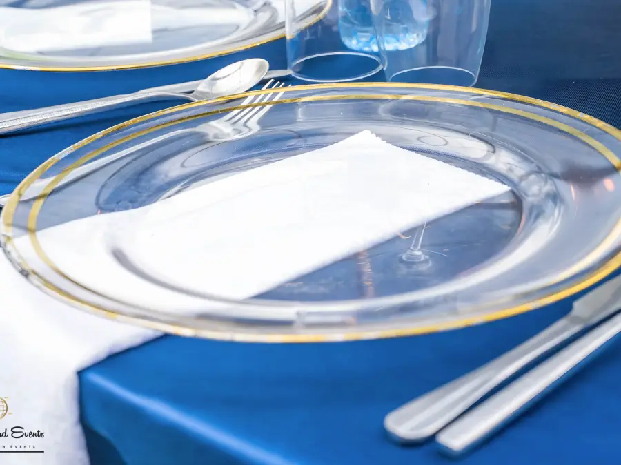 Close-up of a table setting featuring a gold-rimmed clear glass plate, white napkin, and cutlery on a navy blue tablecloth.