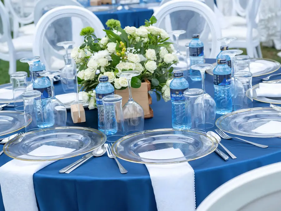 Low angle view of a wedding reception table setting with a white floral centerpiece in a wooden crate, blue tablecloth, and white round-back chairs.