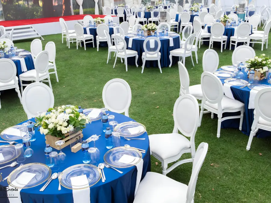Wide shot of an outdoor event reception on a lawn featuring round tables with navy blue tablecloths, white chairs, and simple floral centerpieces.