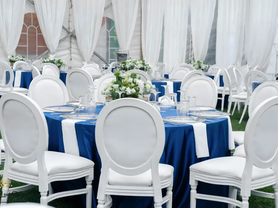 Wide interior shot of a white event tent with draped ceilings, blue round tables, white Louis chairs, and floral centerpieces.
