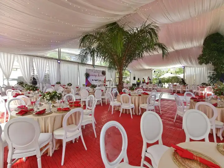 Interior of a white tent with a red tiled floor, beige round tables, white Louis chairs, and a