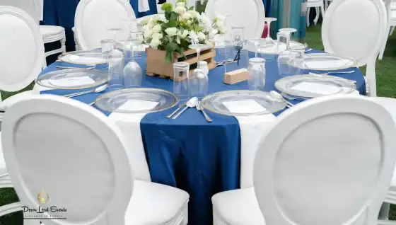 Medium shot of a round table setup with navy blue tablecloth and white round-back chairs, featuring